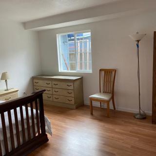 A tidy bedroom with a wooden floor, featuring a dresser, chair, bed, and standing lamp near a window.