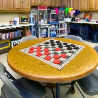 A game room featuring a round table set up for a checkers match with shelves of toys and games in the background.