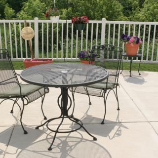 A sunny patio with a glass-top table and metal chairs, surrounded by potted plants and flowers.