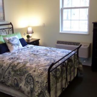 A cozy bedroom with a neatly made bed, a nightstand with a lamp, and a window letting in natural light.