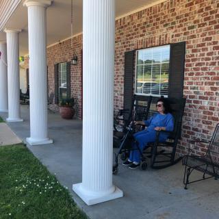 A person in blue attire relaxes on a rocking chair on a porch with large white columns and a brick facade.