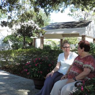 Two people sitting on a bench in a lush garden enjoying a conversation under the shade of trees.