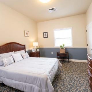 A cozy bedroom with a large bed, nightstand, lamp, and dresser, featuring a window allowing natural light to enter.