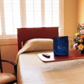 A hospital room with a neatly made bed, featuring a tray holding a remote and gift bag, beside a bouquet of flowers by the window.