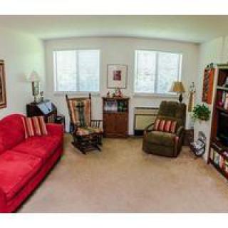 A cozy living room with a red sofa, armchair, and rocking chair, featuring a bookshelf and windows allowing natural light to fill the space.