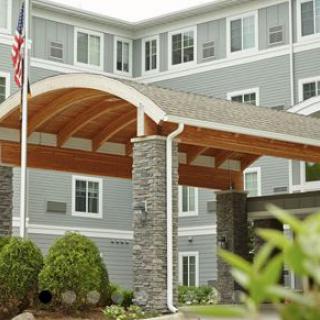 A modern building entrance with stone pillars and a wooden arch, featuring a flag and landscaped greenery.
