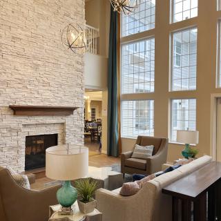 A modern living room with high ceilings, large windows, and a stone fireplace, featuring elegant furniture and a stylish chandelier.