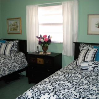 A cozy bedroom featuring two twin beds with patterned bedding, a dark wooden nightstand, and a floral arrangement near the window.