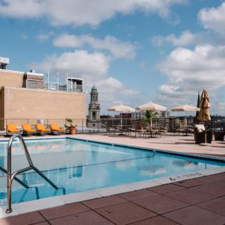 A rooftop swimming pool with deck chairs overlooking the city skyline under a partly cloudy sky.