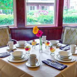 A beautifully set dining table with cups, bowls, and floral centerpieces by a large window.