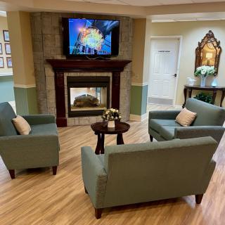 A cozy living room with green sofas arranged around a central table, featuring a fireplace and a wall-mounted television.