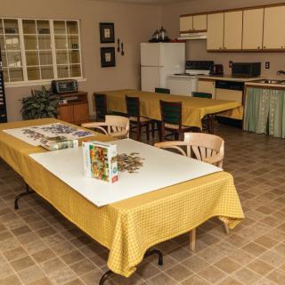 A spacious kitchen and dining area with tables covered in yellow tablecloths, chairs, a refrigerator, and kitchen cabinets.