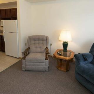 A cozy living room with a gray armchair, a wooden side table with a lamp, and a blue sofa. The kitchen area is visible in the background.