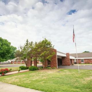 A brick building with an entrance, surrounded by greenery and a flagpole, under a cloudy sky.