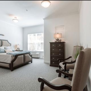 A spacious and elegantly furnished bedroom featuring a bed, dresser, chairs, and a large window allowing natural light.