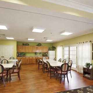 A spacious dining room with multiple tables and chairs, adjacent to a small kitchen area, featuring hardwood flooring and large windows providing plenty of natural light.