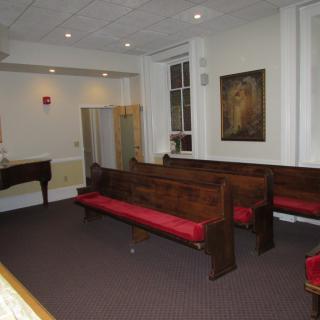 A small church interior with wooden pews, a piano, and a painting on the wall.
