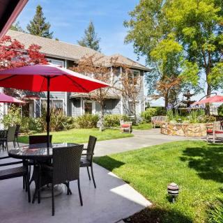 A sunny outdoor patio area with tables, chairs, and red umbrellas surrounded by a lush garden and trees.