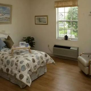 A cozy bedroom with a neatly made bed, wooden flooring, and natural light coming through the window.