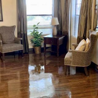A cozy living room with two chairs, a wooden table, and a lamp placed near a window with curtains. A plant adds a touch of greenery on the polished hardwood floor.