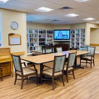 A cozy library space with a large table, surrounded by chairs, bookshelves, and a television.