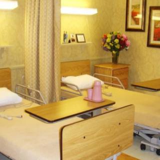 A hospital room featuring two patient beds with adjustable tables and a floral arrangement on a side table.