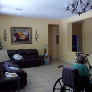 A cozy living room with a person in a wheelchair, featuring a sofa, painting, and chandelier.