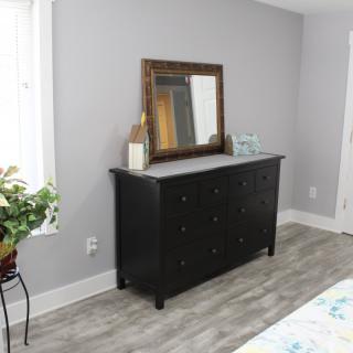 A bedroom interior featuring a dark dresser with a mirror above, decorated with plants and accessories.