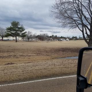 A cloudy day view from a vehicle, showcasing a stretch of road lined with trees and open fields.
