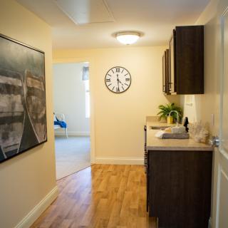 A well-lit hallway with wooden flooring, featuring a wall-mounted clock and artwork, along with dark cabinets for storage.