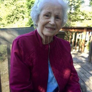 An elderly woman smiles while sitting outdoors in a red jacket.