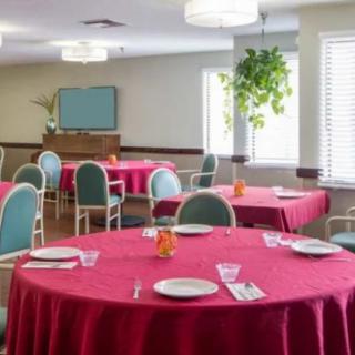 A dining area with tables covered in red tablecloths, surrounded by teal chairs, and accented by natural light from the windows.