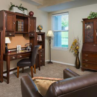 A cozy home office setup with a wooden desk, leather chair, and bookshelf, illuminated by natural light from the window.