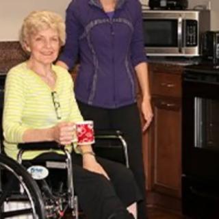 An elderly woman in a wheelchair holding a mug, with a caregiver standing beside her in the kitchen.
