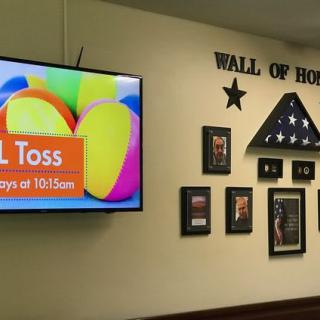 A 'Wall of Honor' display featuring framed photos and a folded flag, with a television advertising a ball toss event.
