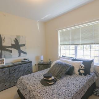 A cozy bedroom with a neatly made bed, decorative pillows, a dresser, and a large window letting in natural light.