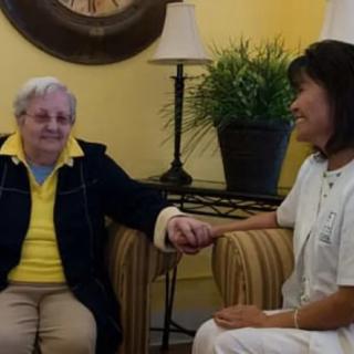 An elderly person and a caregiver share a moment while holding hands indoors.