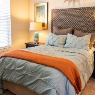 A cozy bedroom with a neatly made bed featuring blue and orange bedding, complemented by a bedside lamp and artwork on the wall.