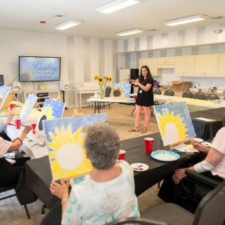 A group of adults participating in a painting workshop, each working on their own canvas while an instructor guides the session.