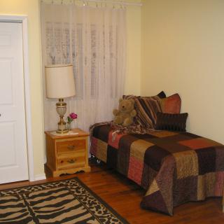 A cozy bedroom corner featuring a small bed with decorative pillows, a teddy bear, a nightstand with a lamp, and a patterned rug.
