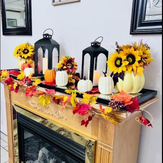 A cozy autumn-themed fireplace decoration with sunflowers, pumpkins, candles, and colorful leaves.