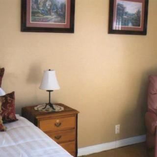 A cozy bedroom corner with a nightstand, lamp, artwork, and a comfortable chair.
