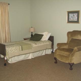 A simple bedroom with a single bed, an armchair, and a lamp beside the bed.