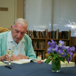 An elderly author signs a book in a library setting with flowers on the table.