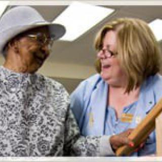 A caregiver assists an elderly woman, both smiling warmly at each other.