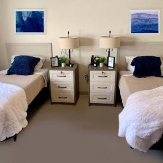 A cozy and minimalist bedroom featuring twin beds with blue pillows, matching nightstands, and calming artwork on the walls.