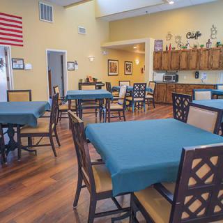 A spacious dining room with wooden floors, tables and chairs neatly arranged, and a kitchen area in the background.