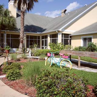 A house with a landscaped garden featuring a decorative bicycle and a palm tree on a sunny day.