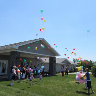 A group of people participate in a balloon release on a sunny day outside a house.