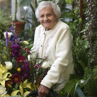 An elderly woman smiling in a lush indoor garden filled with colorful flowers.